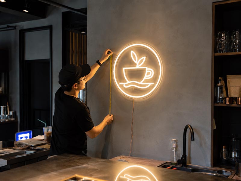 Installer measuring a custom LED neon logo sign on a cafe wall to check size, brightness, and placement before production approval
