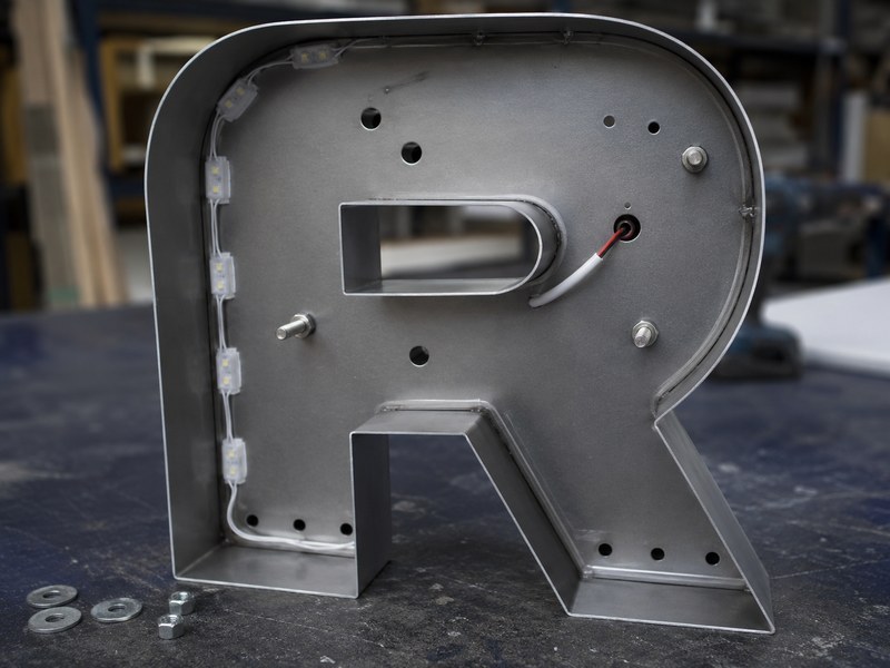 Rear view of a channel letter showing mounting holes, threaded studs, wire exits, and drainage openings
