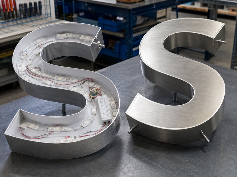 Open illuminated channel letter with LED components beside a brushed non-illuminated metal letter